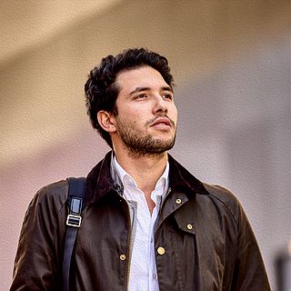Young man with brown jacket and shoulder bag in front of Messe Basel – modern, natural look captured by the Bluesun photography team.