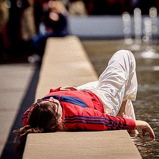 Woman in red track jacket relaxing by the fountain in front of Art Basel – a sunny moment of rest captured by the Bluesun photography team.