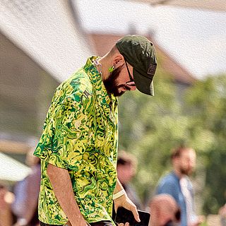 Man in a green patterned shirt, baseball cap and sunglasses walking casually with a smartphone in hand in front of Messe Basel – captured by Bluesun Event Photography.