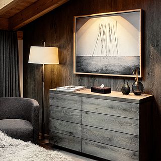 Designer wooden chest of drawers, floor lamp and framed artwork in a stylish room with dark wall paneling - Bluesun Interior Photography.